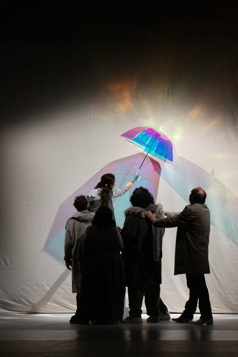 A group of performers gather beneath a brightly lit umbrella on a stage.