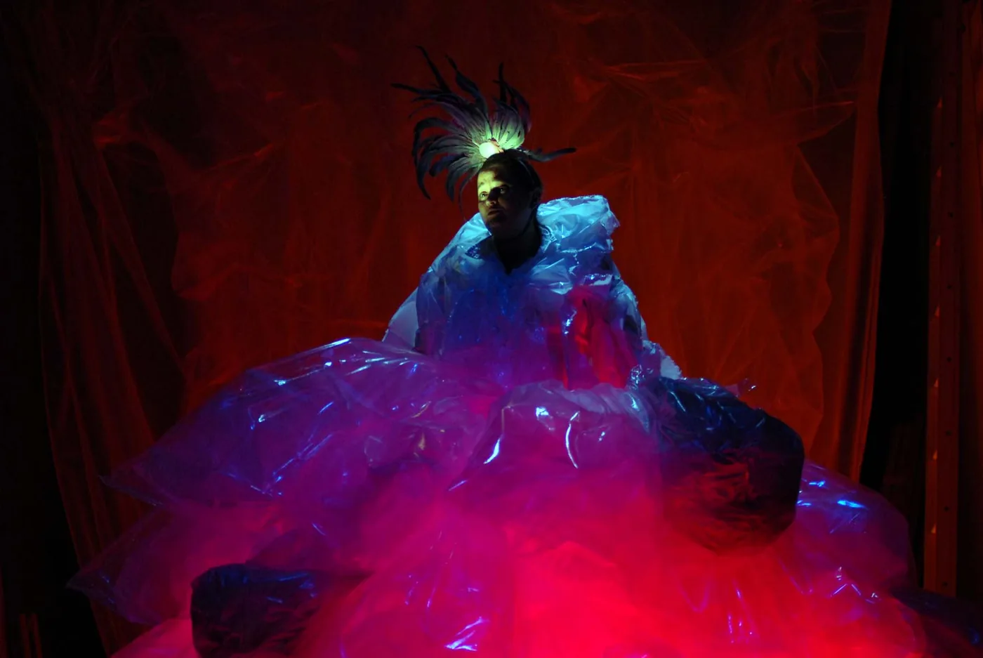 A performer in a glowing translucent costume sits against a red stage backdrop.