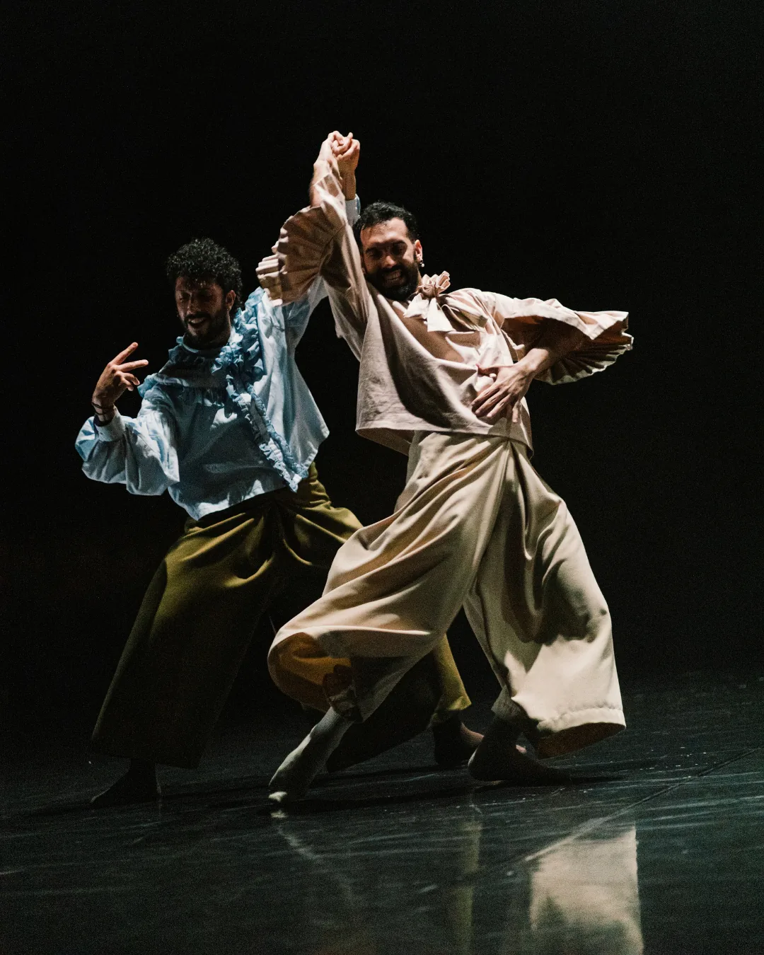 Two performers in flowing costumes dance under a spotlight on a dark stage.