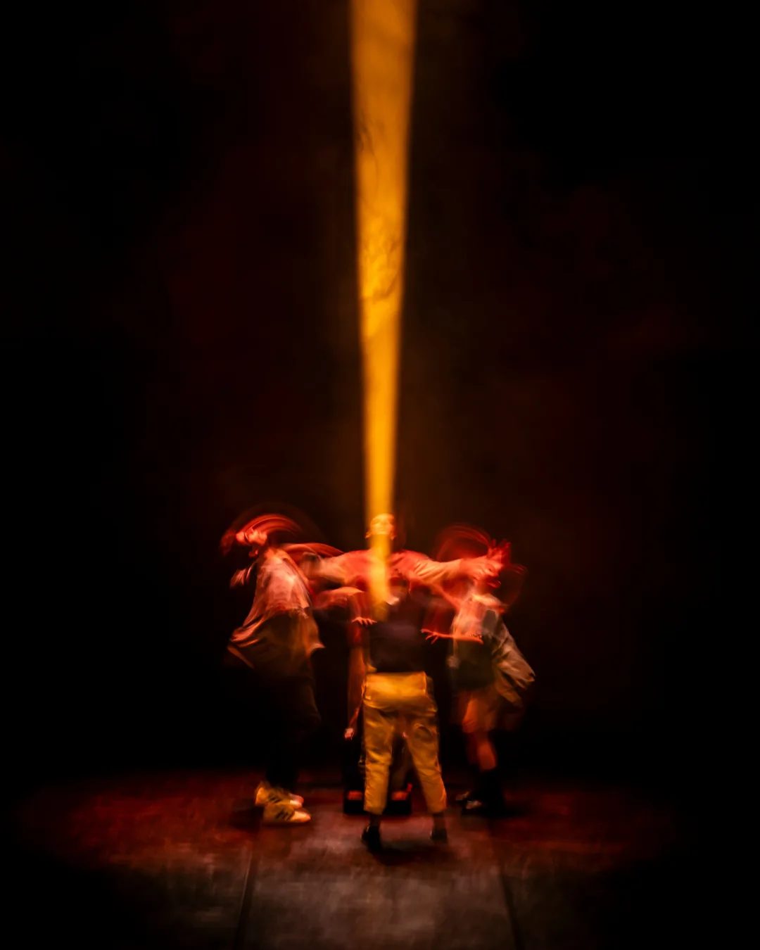 Several performers move beneath a narrow amber spotlight on a dark stage.