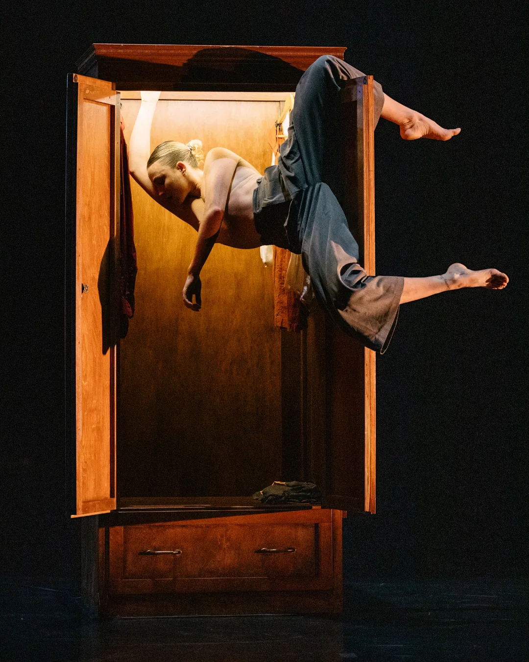 A dancer leans out of an open wooden cabinet under warm stage lighting.