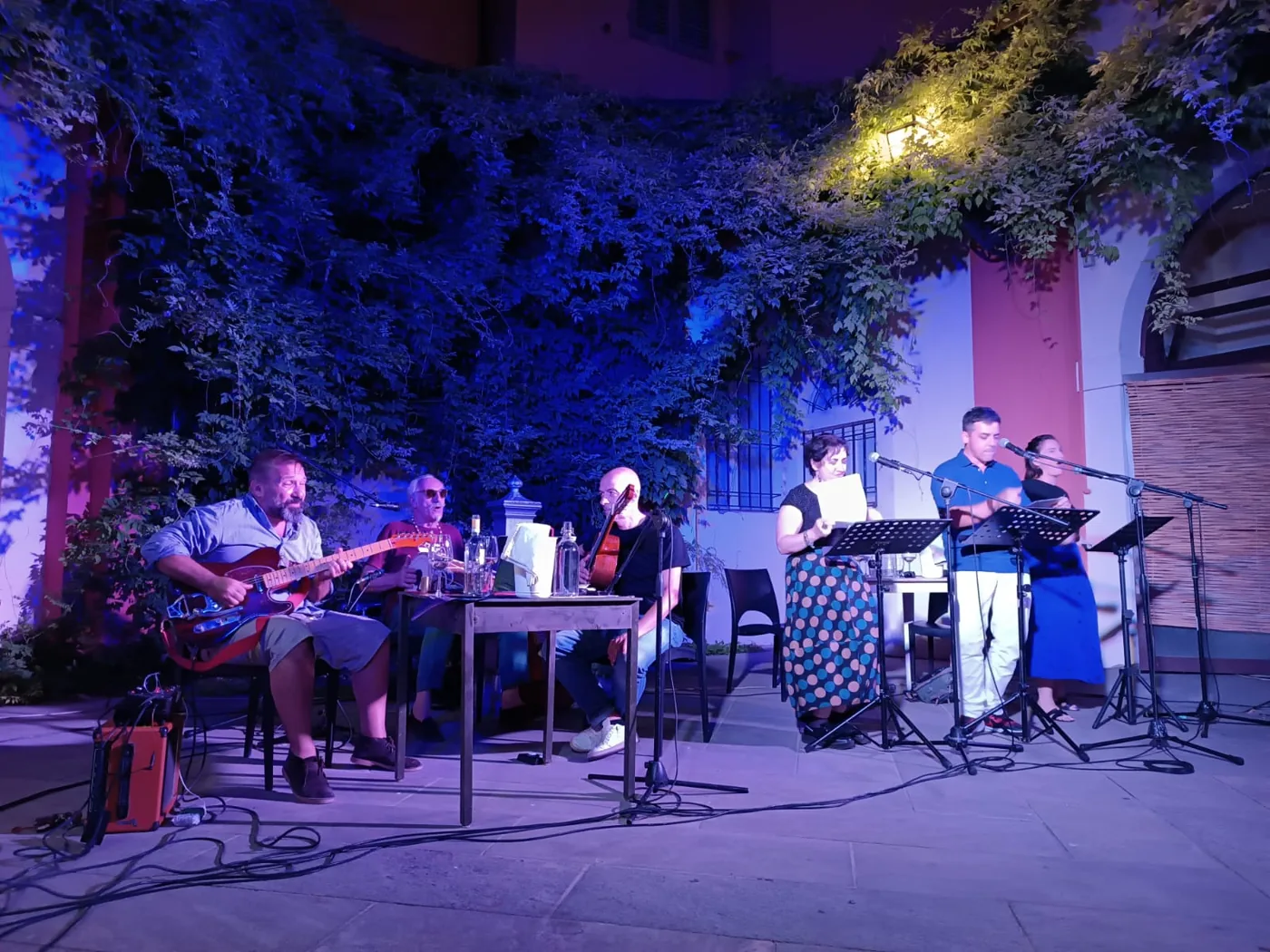 People read from pages at microphones while musicians perform in an outdoor courtyard.