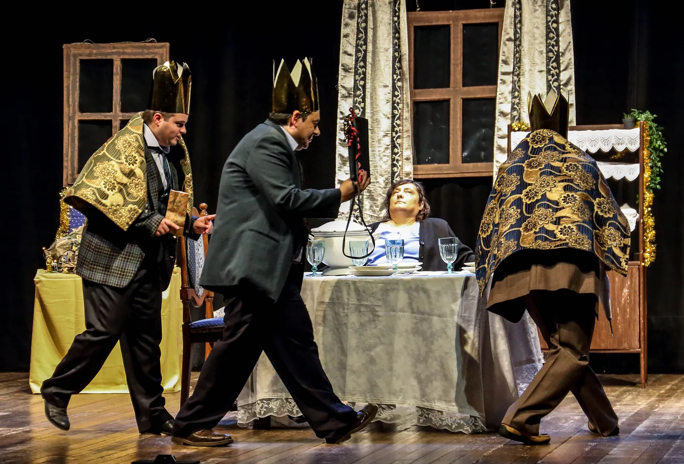 Two costumed performers approach a seated figure at a table on stage.