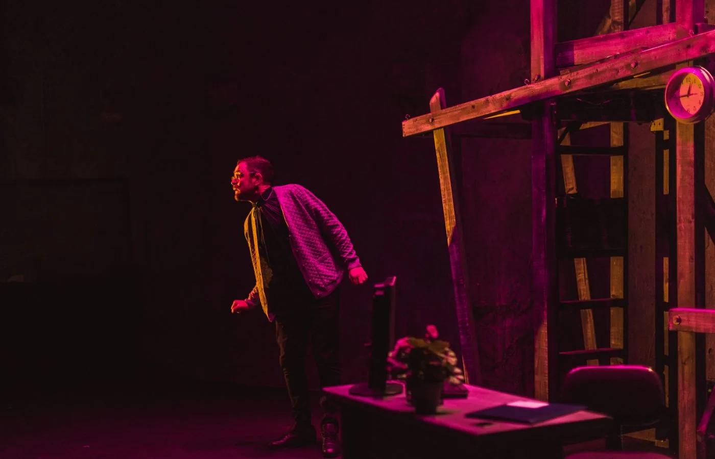 A performer stands beside a wooden stage set under magenta lighting.