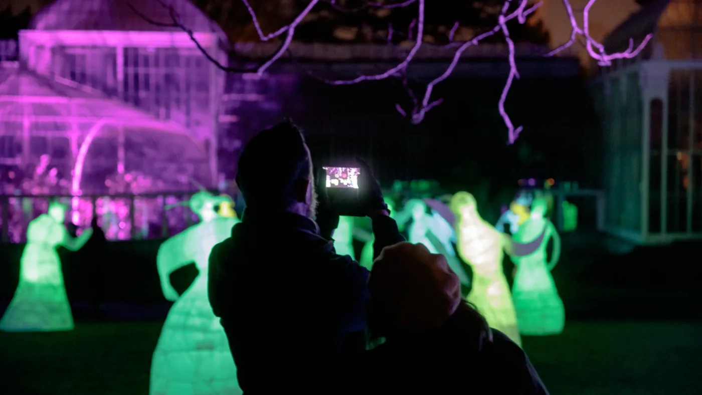 A person photographs illuminated figures in a garden at night.