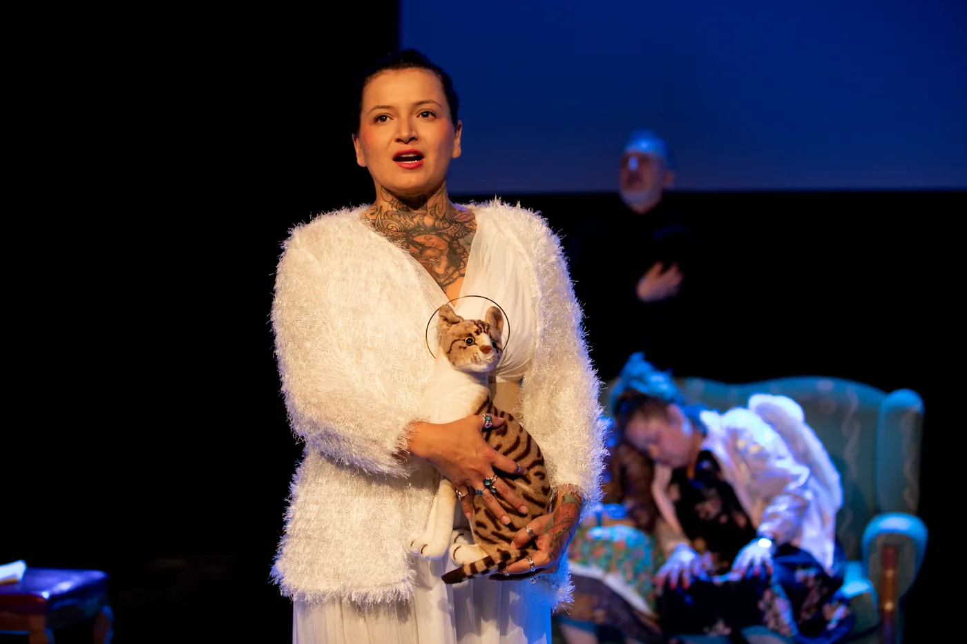 A performer in a white costume holds a cat-shaped prop on a blue-lit stage.