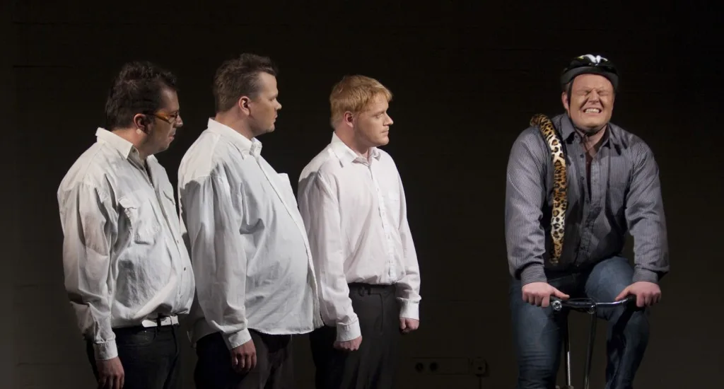 Four performers stand on a dark stage; one holds bicycle handlebars.