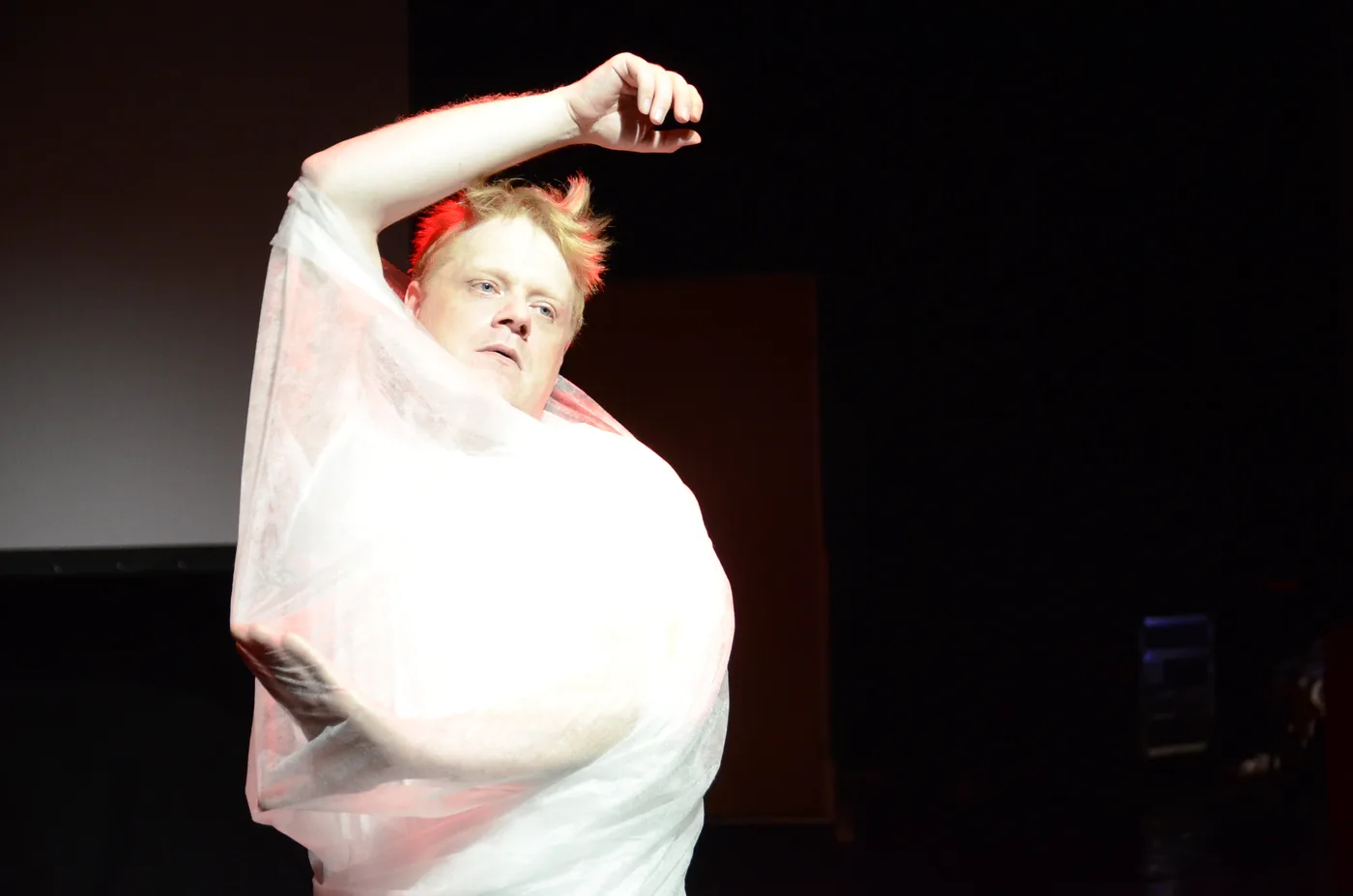A performer in white fabric raises one arm under a spotlight.
